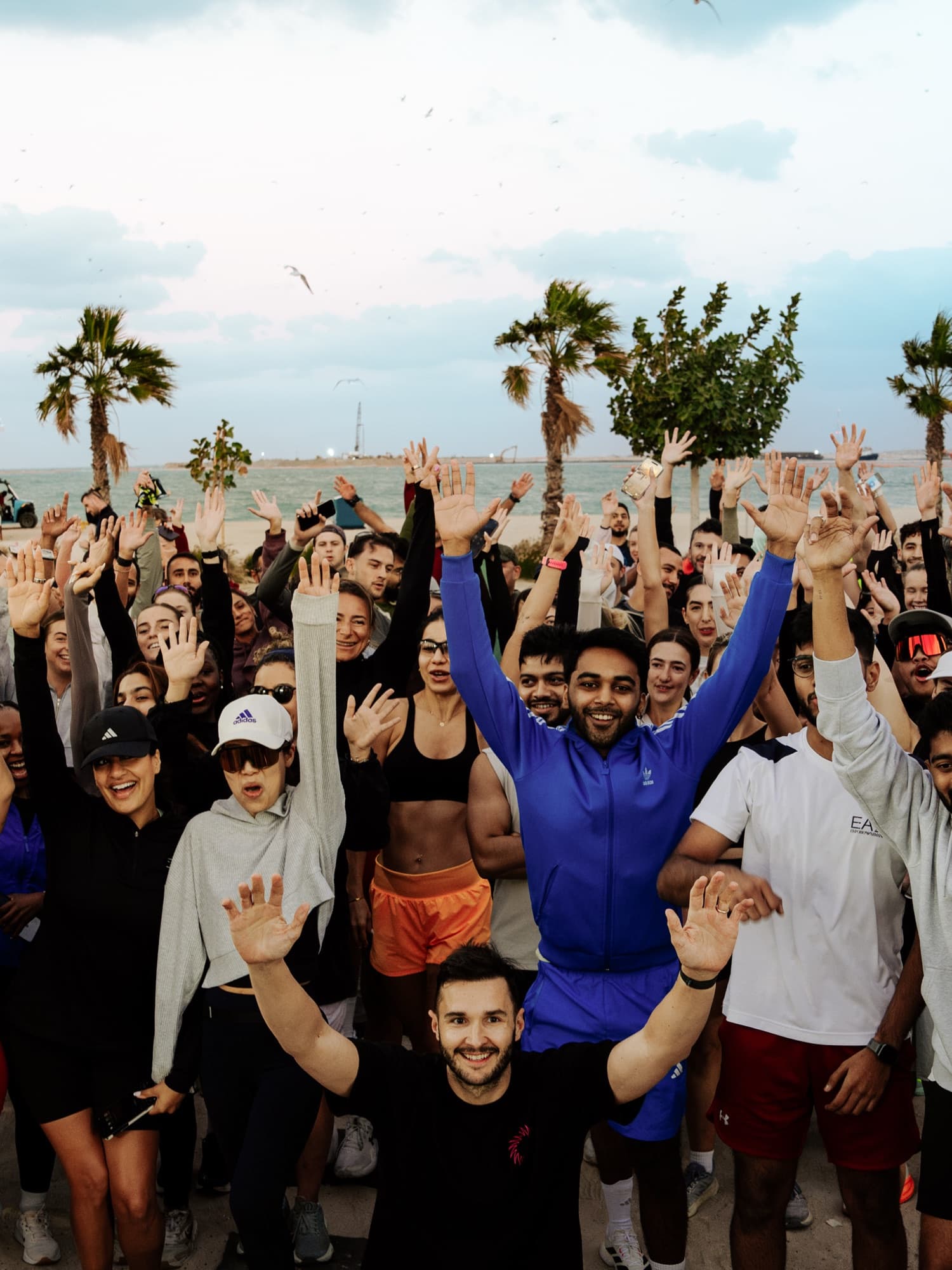 A large run club crowd cheering with arms raised on the beachfront, palms and the Gulf behind